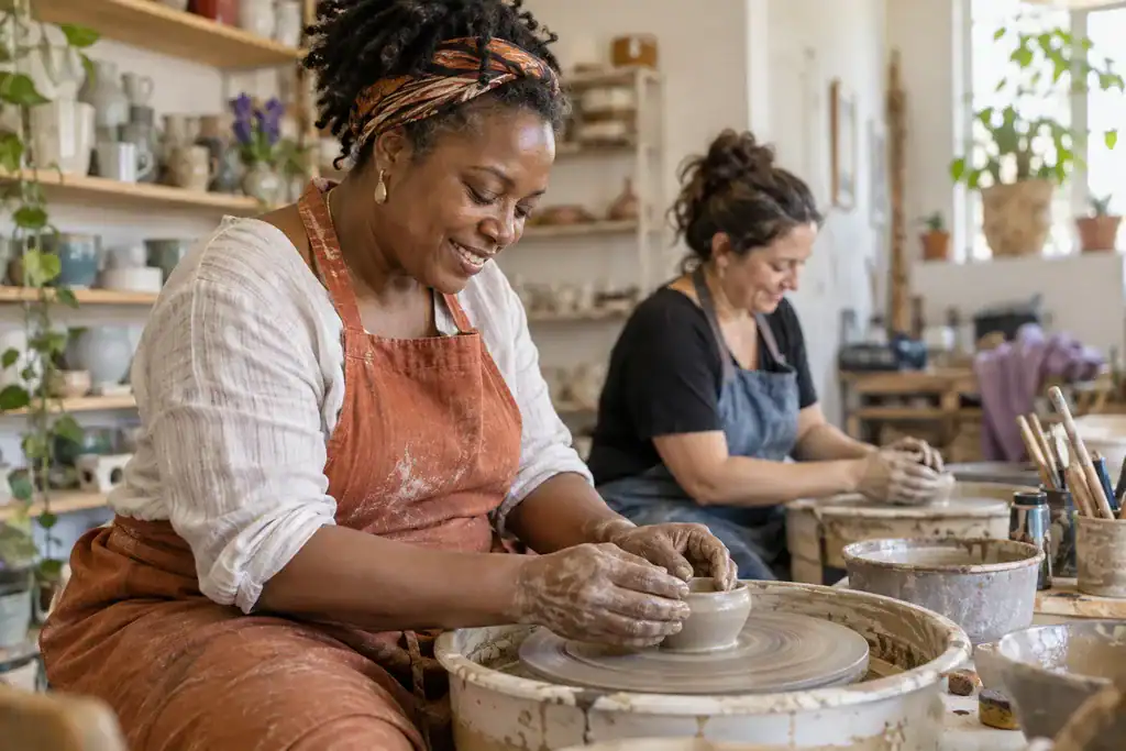 Two women shaping clay bowls at pottery wheels in a sunlit ceramics studio.